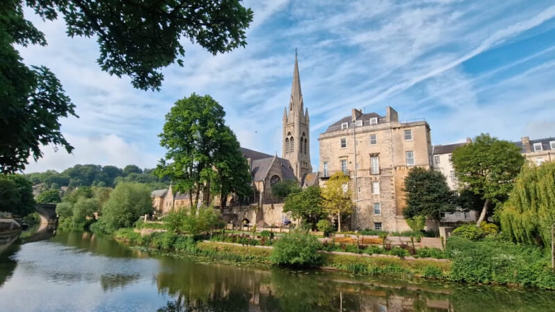 Bath, England, featuring historic stone buildings, a church with a tall spire, and lush greenery reflected in a river