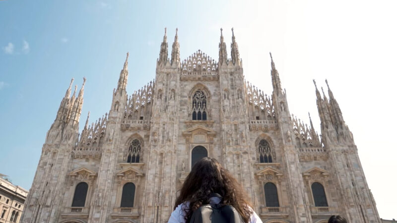 Person with a backpack stands in front of the Milan Cathedral (Duomo di Milano), gazing up at its intricate Gothic architecture